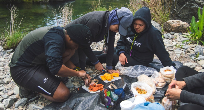 a group of people prepare a meal on a shore beside a body of water on an outward bound course for bipoc